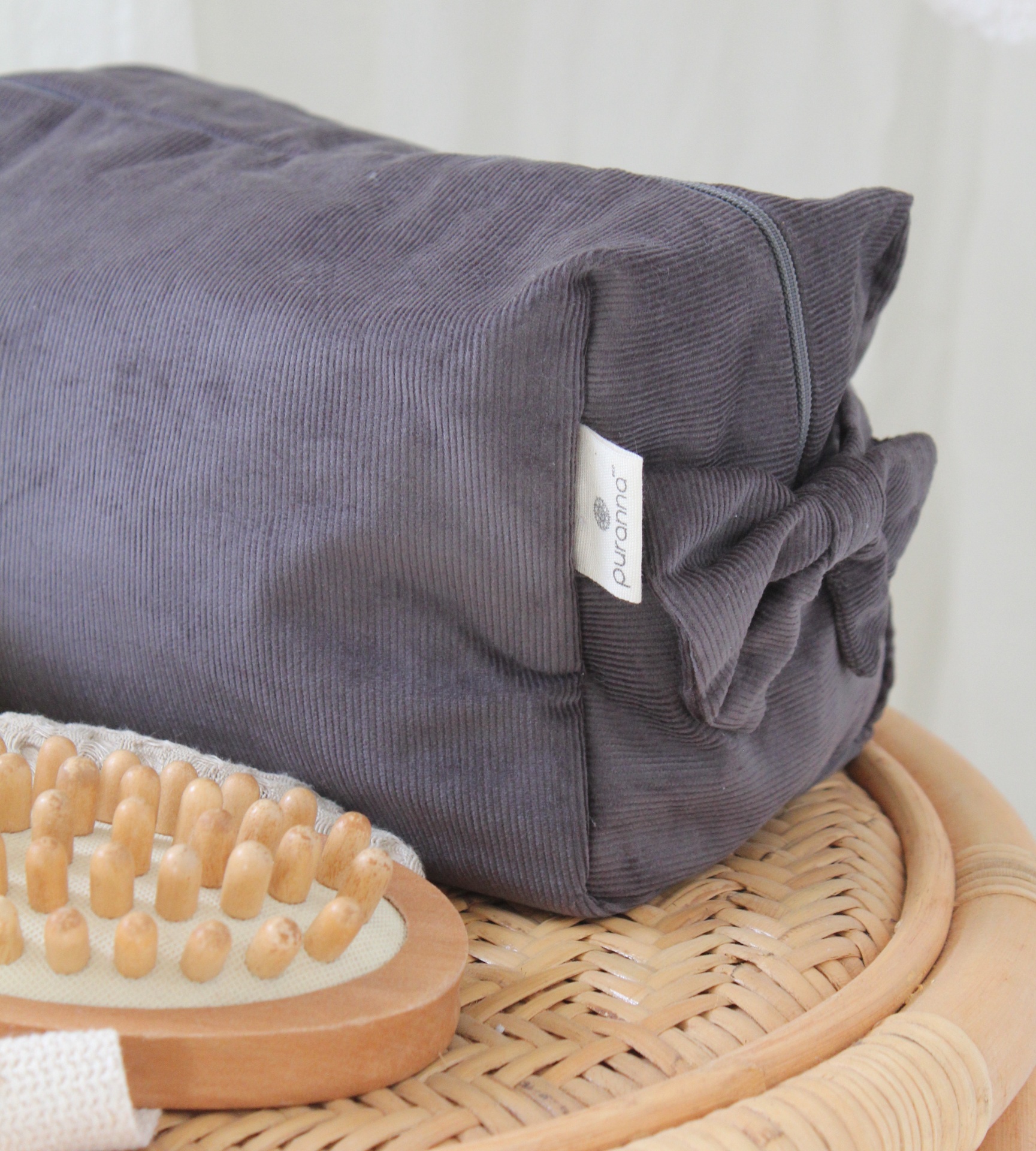 Almofada cinzenta escura com etiqueta Puravida sobre rattan, ao lado de escova de madeira