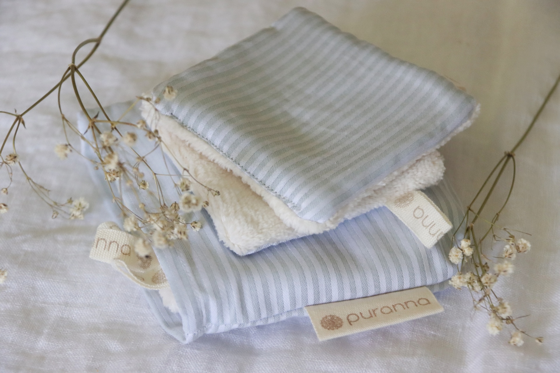 Três toalhetes macios em azul claro, branco e creme com etiqueta puranna e pequenos ramos florais secos