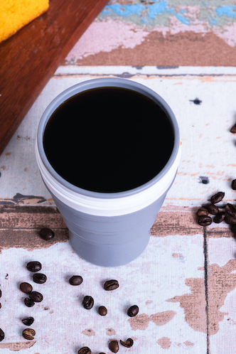 Caneca cinzenta com café preto e grãos de café ao redor sobre superfície manchada