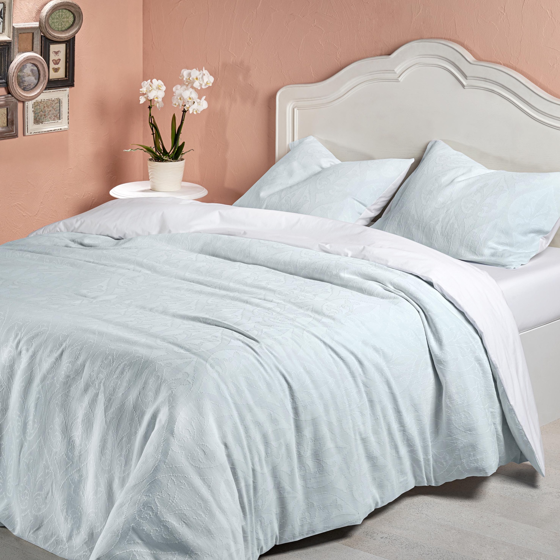 Cama de casal com roupa de cama azul clara e cabeceira branca, mesa com vaso de flores brancas e parede cor de rosa