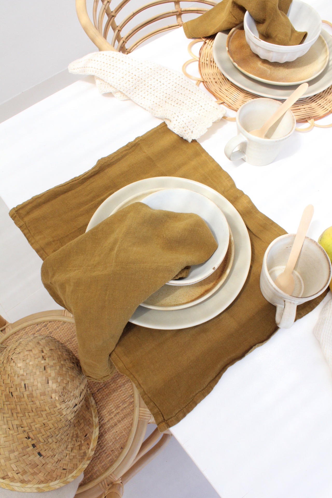 Mesa posta com utensílios de cerâmica branca e castanha, toalhete e guardanapo castanhos, cadeira de rattan com chapéu de palha.