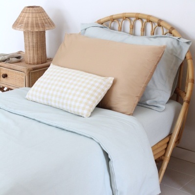 Cama com estrutura em rattan, roupa de cama azul claro, almofadas bege e xadrez amarelo e branco, e candeeiro em rattan numa mesa de cabeceira