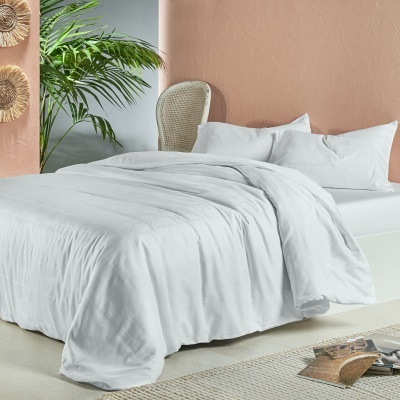 Cama de casal com roupa de cama branca, almofadas, edredão, cadeira de rattan e planta verde próximo a parede salmão.
