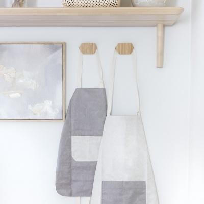 Dois aventais pendurados em gancho na parede com um quadro abstrato ao lado e prateleira de madeira acima