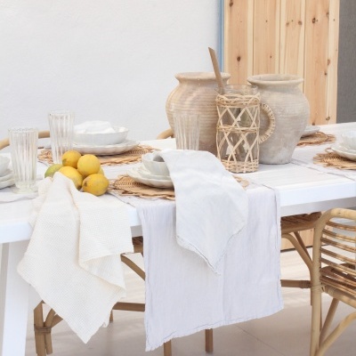 Mesa branca posta com toalhas de linho, frutas amarelas, vasos de cerâmica e abajures de vime em ambiente branco e madeira clara