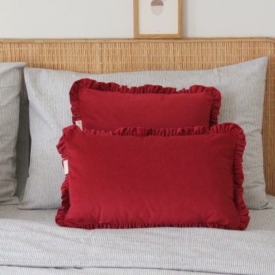 Duas almofadas vermelhas com folhos numa cama com roupa de cama cinzenta riscada e cabeceira em rattan.