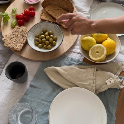 Mesa posta com pão, azeitonas, tomates, limões e azeite sobre tábua redonda de madeira, pratos e copos sobre toalhas de mesa coloridas.