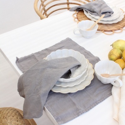 Mesa branca com conjunto de loiça branca ondulada, panos cinzentos e cadeiras de rattan