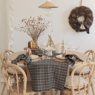 Mesa redonda de madeira com toalha xadrez e decoração de flores secas, cadeiras de vime, guirlanda na parede e lâmpada de palha