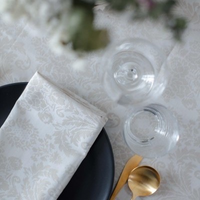 Mesa com toalha branca floral, prato preto, guardanapo branco, talheres dourados e copos transparentes