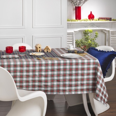 Mesa com toalha xadrez, castiçais, miniaturas de casas, cadeiras brancas e almofada azul num interior com lareira branca e vasos vermelhos