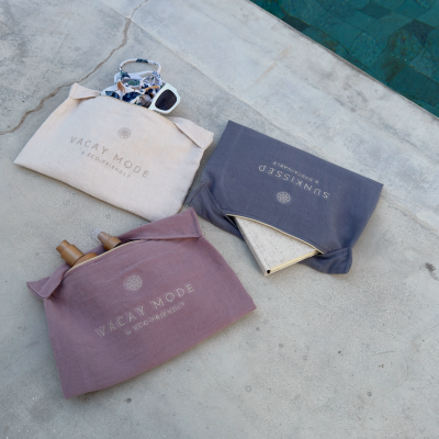 Três sacos de tecido com fecho e textos VACAY MODE e SUNKISSED sobre cimento junto a piscina