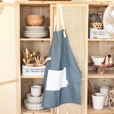 Avental azul com bolso branco pendurado numa porta de armário de cozinha de madeira com pratos e utensílios.