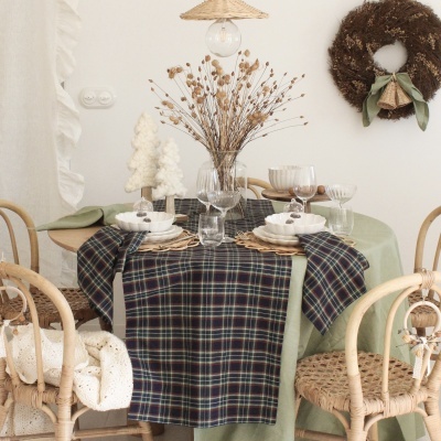 Mesa de jantar decorada com toalha verde e azul xadrez, vasos com flores secas, pratos e copos, rodeada por cadeiras de madeira e coroa de flores na parede