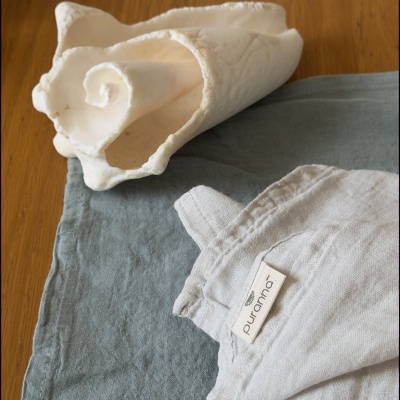 Mesa com guardanapo cinzento-azulado, toalha de linho cinzento clara com etiqueta Puranos, concha decorativa branca e copo transparente