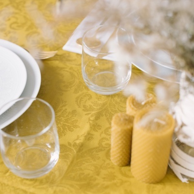 Mesa com toalha amarela, copos de vidro, pratos brancos, velas amarelas e vaso com flores secas.