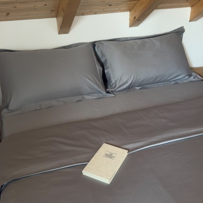 Roupa de cama cinza com duas fronhas e um livro sobre um lençol esticado numa cama de madeira