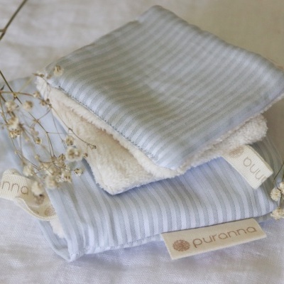 Três toalhetes macios em azul claro, branco e creme com etiqueta puranna e pequenos ramos florais secos