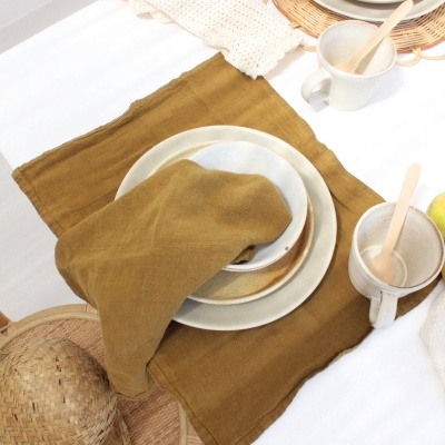 Mesa posta com utensílios de cerâmica branca e castanha, toalhete e guardanapo castanhos, cadeira de rattan com chapéu de palha.