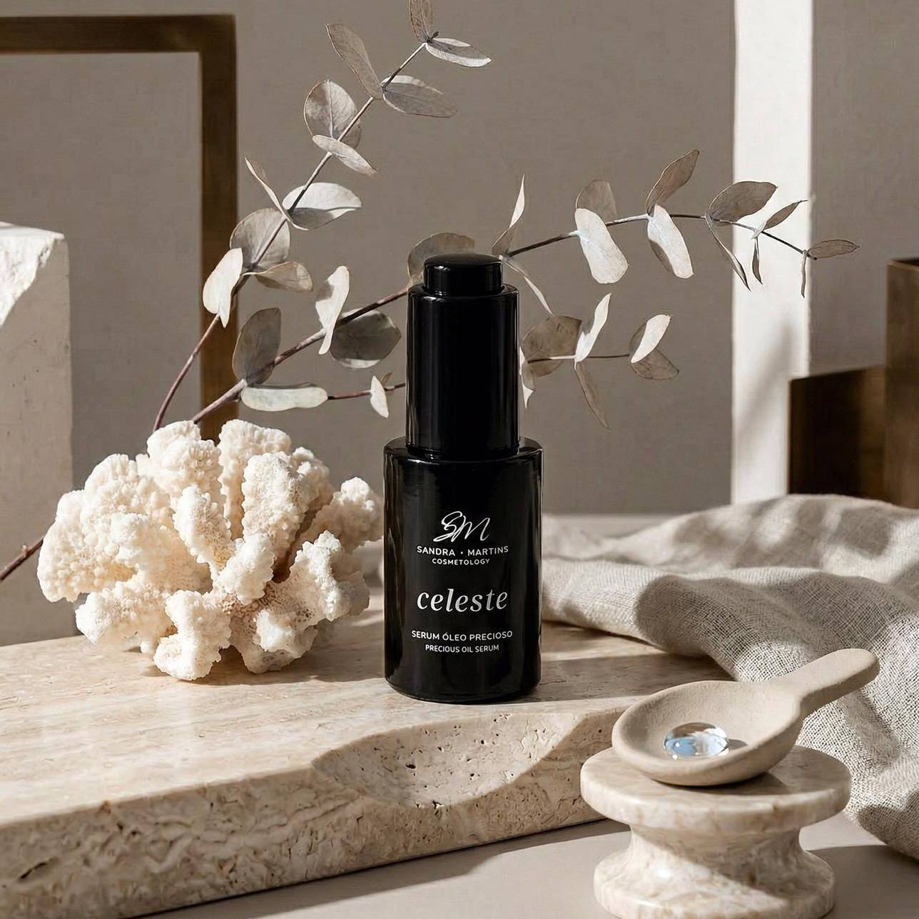 Sérum Óleo Precioso Celeste Frasco preto de sérum com coral branco e ramo seco sobre pedra clara.