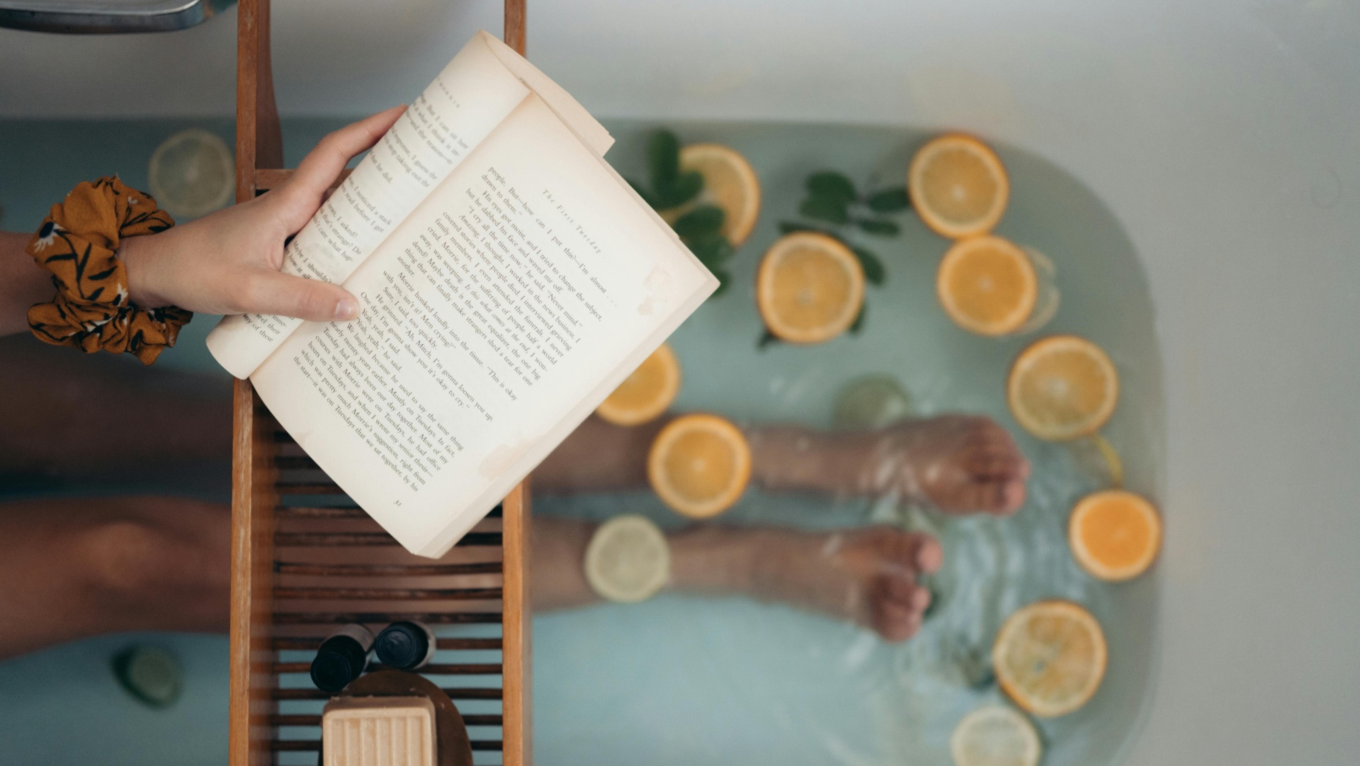 Banheira com água, limões, folhas, pessoa a ler livro e bancada de madeira