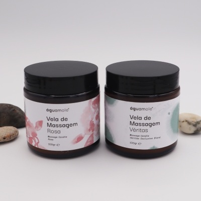 Dois frascos de velas de massagem ág u amole com rótulos brancos e tampas pretas, um com aroma rosa e outro Vér itas, sobre fundo branco com pedras pequenas ao redor.