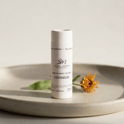 Bálsamo labial branco com texto calendula ao lado de flor amarela seca numa base redonda bege