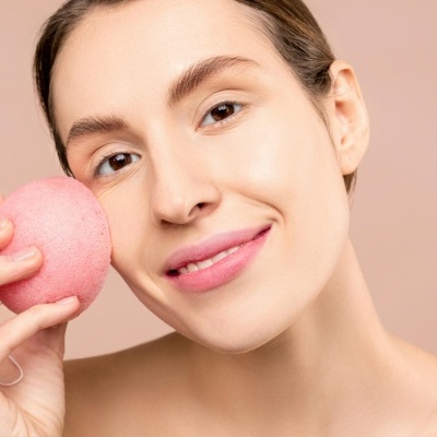 Mulher segurando esponja de limpeza facial rosa junto ao rosto