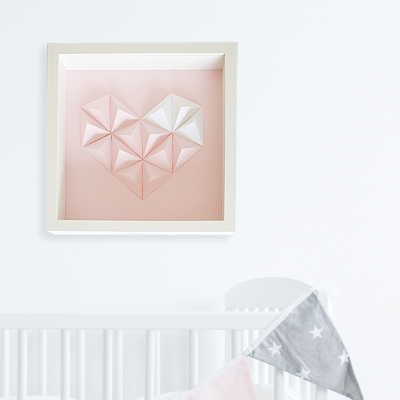 Quadro com coração de papel rosa e branco em moldura branca acima de berço branco com bandeiras cinza e rosa