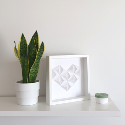 Vaso branco com planta verde, moldura branca com coração geométrico, pequeno recipiente branco com tampa verde numa prateleira branca.