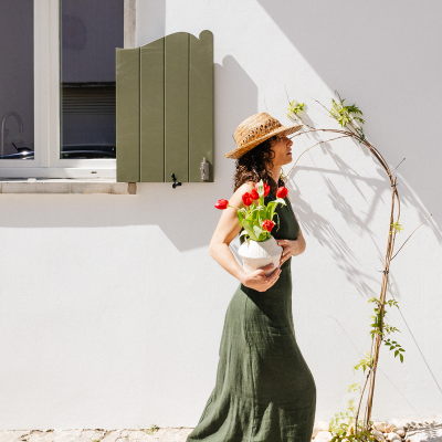 Mulher com vestido verde e chapéu de palha segurando jarra com tulipas vermelhas junto a parede branca com janela e planta trepadeira.