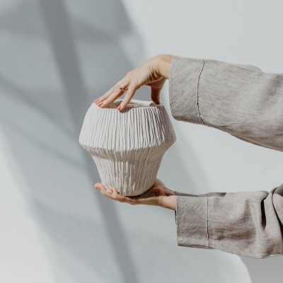 Vaso cerâmico branco com textura e formato arredondado segurado por mãos com mangas cinza