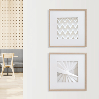 Dois quadros decorativos brancos com padrões geométricos em relevo numa parede branca perto de cadeira e mesa de madeira clara