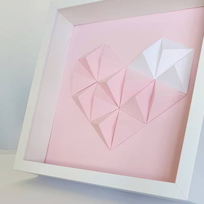 Quadro com molde branco, coração de origami rosa e branco em fundo rosa.
