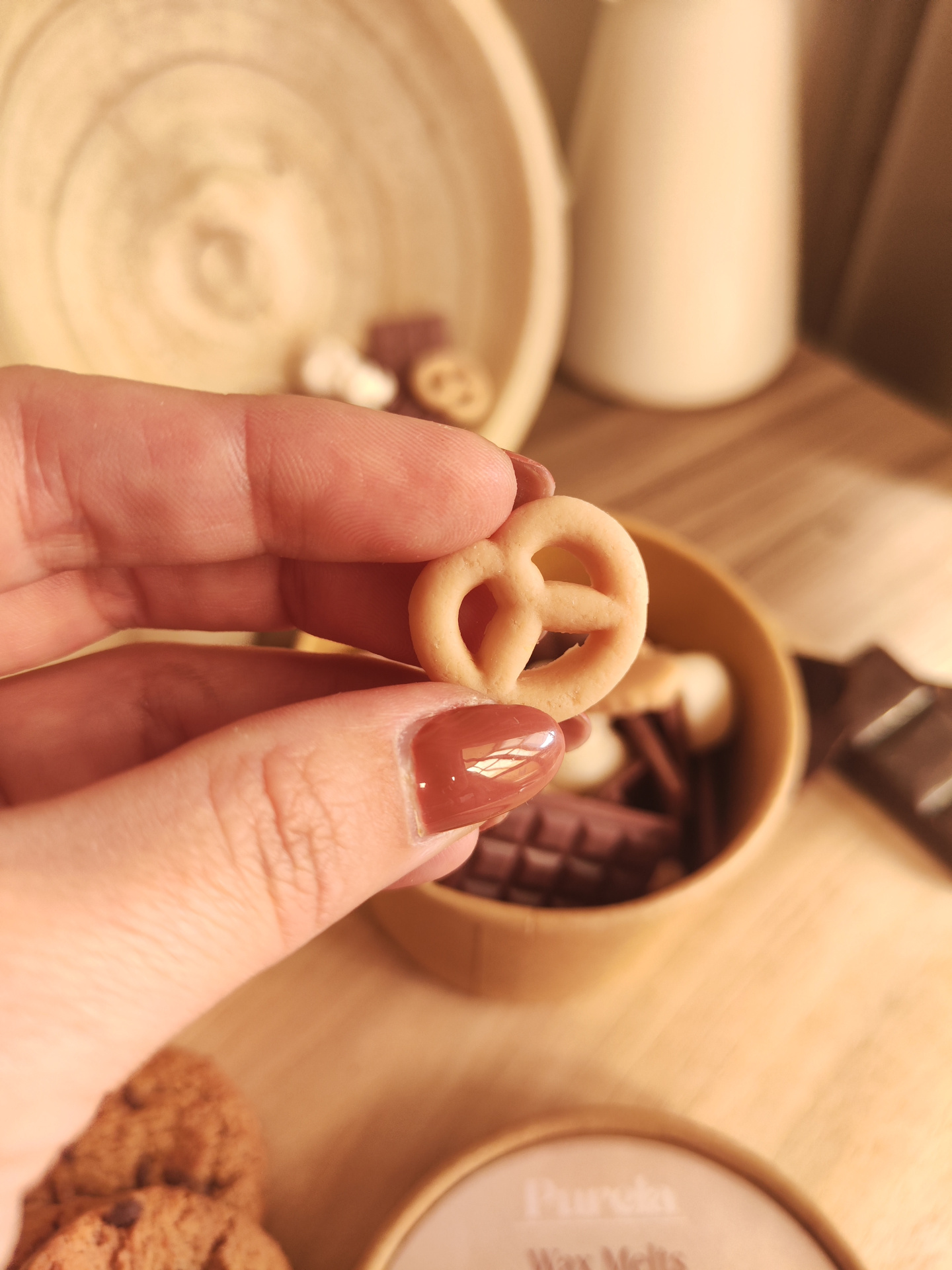 Wax Melt Mimo de Cacau Bolacha em forma de pretzel nas mãos com recipiente de bambu ao fundo