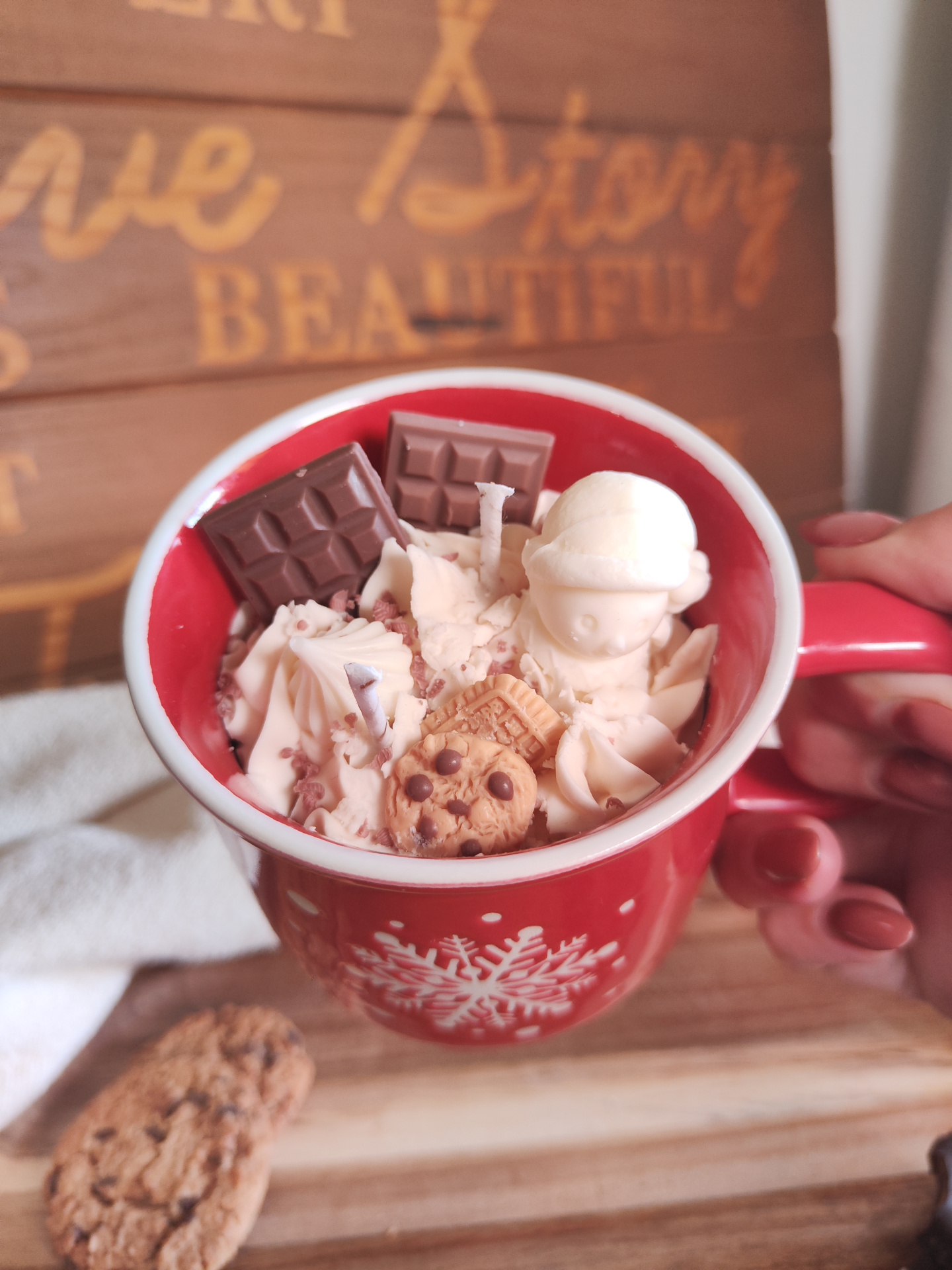 Vela Caneca Mimo de Cacau Caneca vermelha com decoração de floco de neve branco, chantilly e chocolates, sobre tábua de madeira com biscoitos