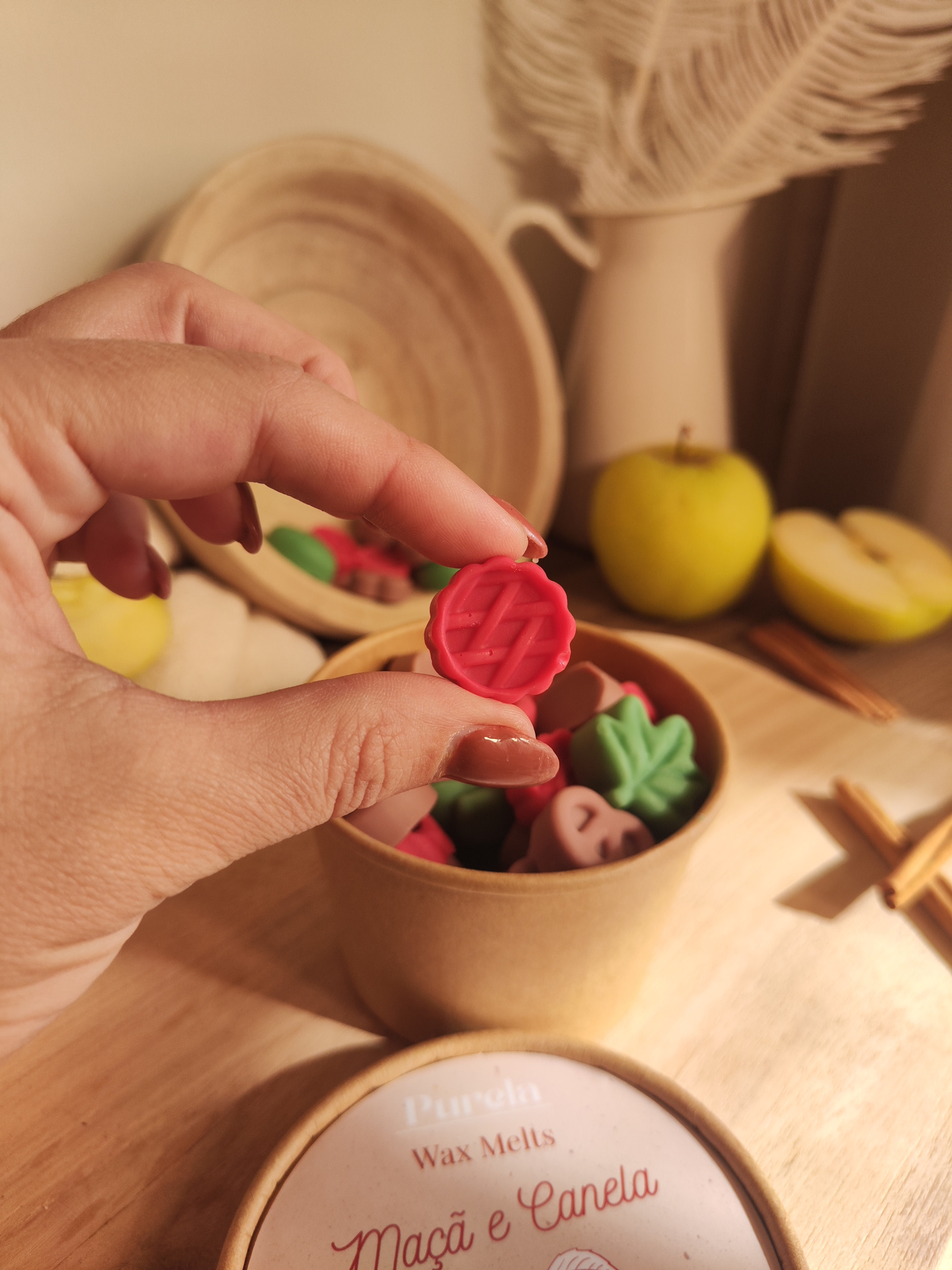 Wax Melt Maçã e Canela Mão segurando cera vermelha decorativa com caixa de madeira, maçã verde e pau de canela ao fundo.