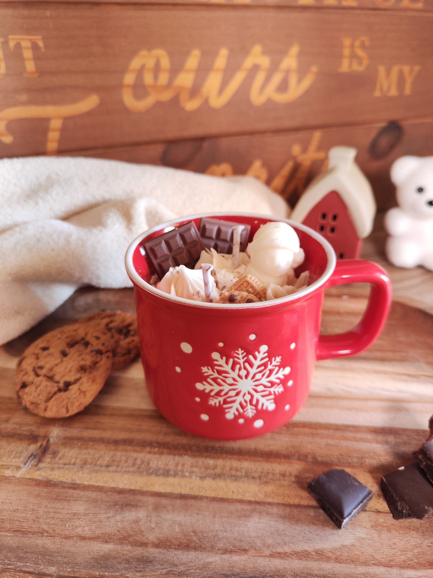 Vela Caneca Mimo de Cacau Caneca vermelha com floco de neve branco cheia de chantilly e chocolates, biscoitos e decoração natalícia ao fundo