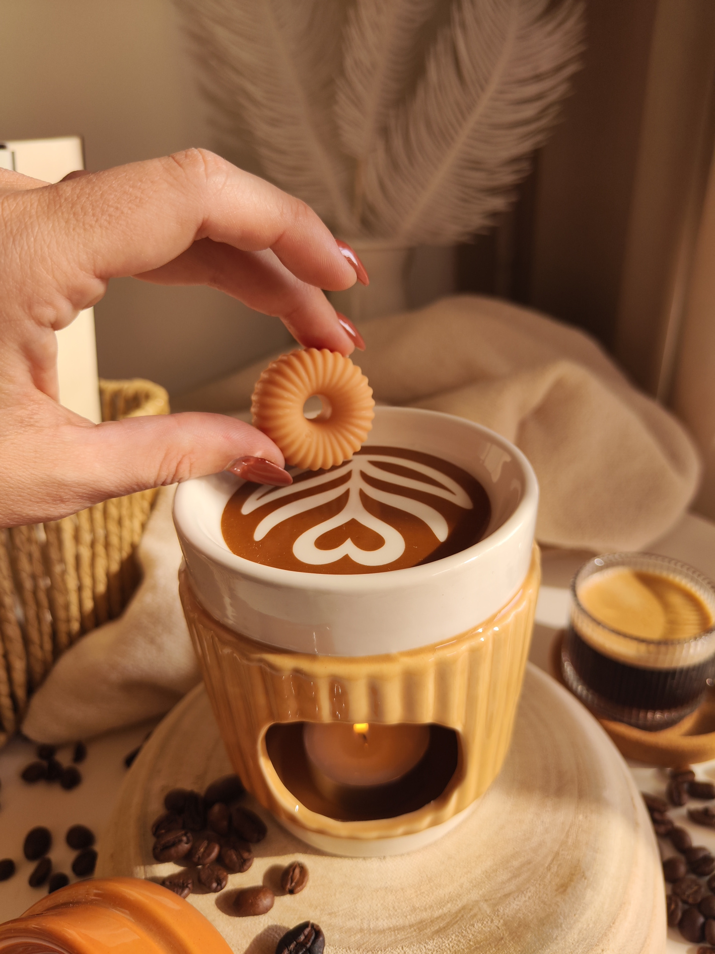 Queimador Copo de café Mão a segurar biscoito sobre chávena com café latte art e grãos de café ao redor.