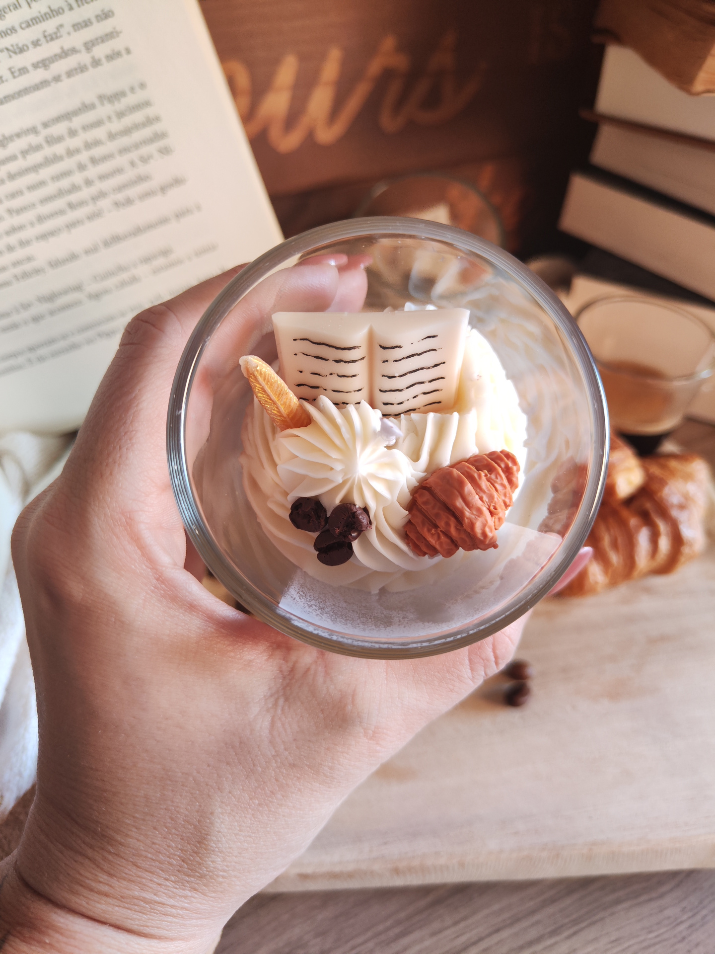 Vela Só mais um Capítulo Sobremesa cremosa num copo decorada com livro de chocolate, pena e pinha, com livros e croissants ao fundo