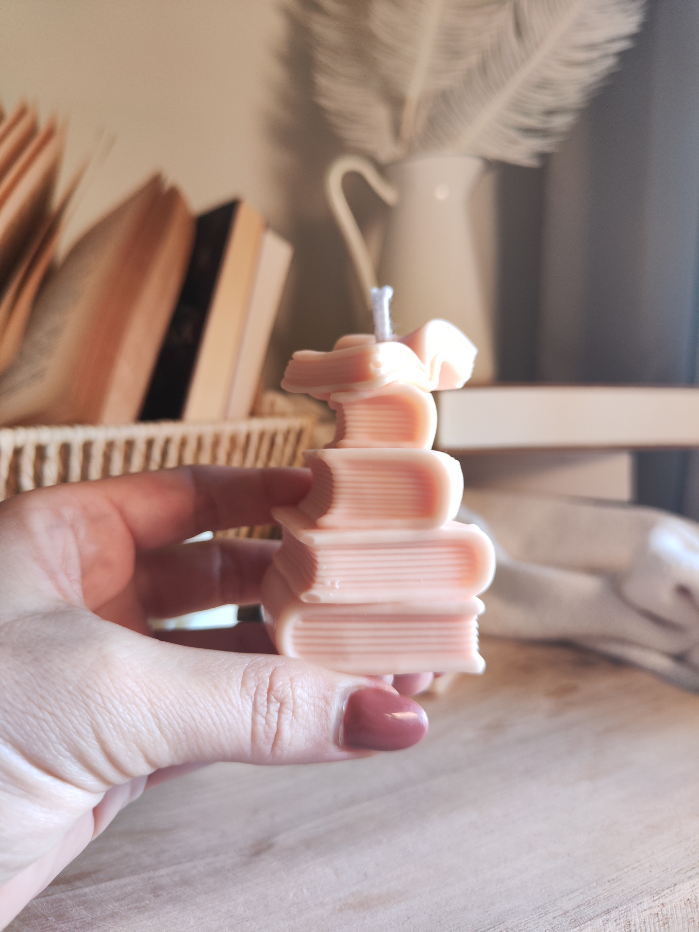 Vela TBR Vela em forma de livros empilhados cor-de-rosa com textura e mão à frente, livros e vaso ao fundo