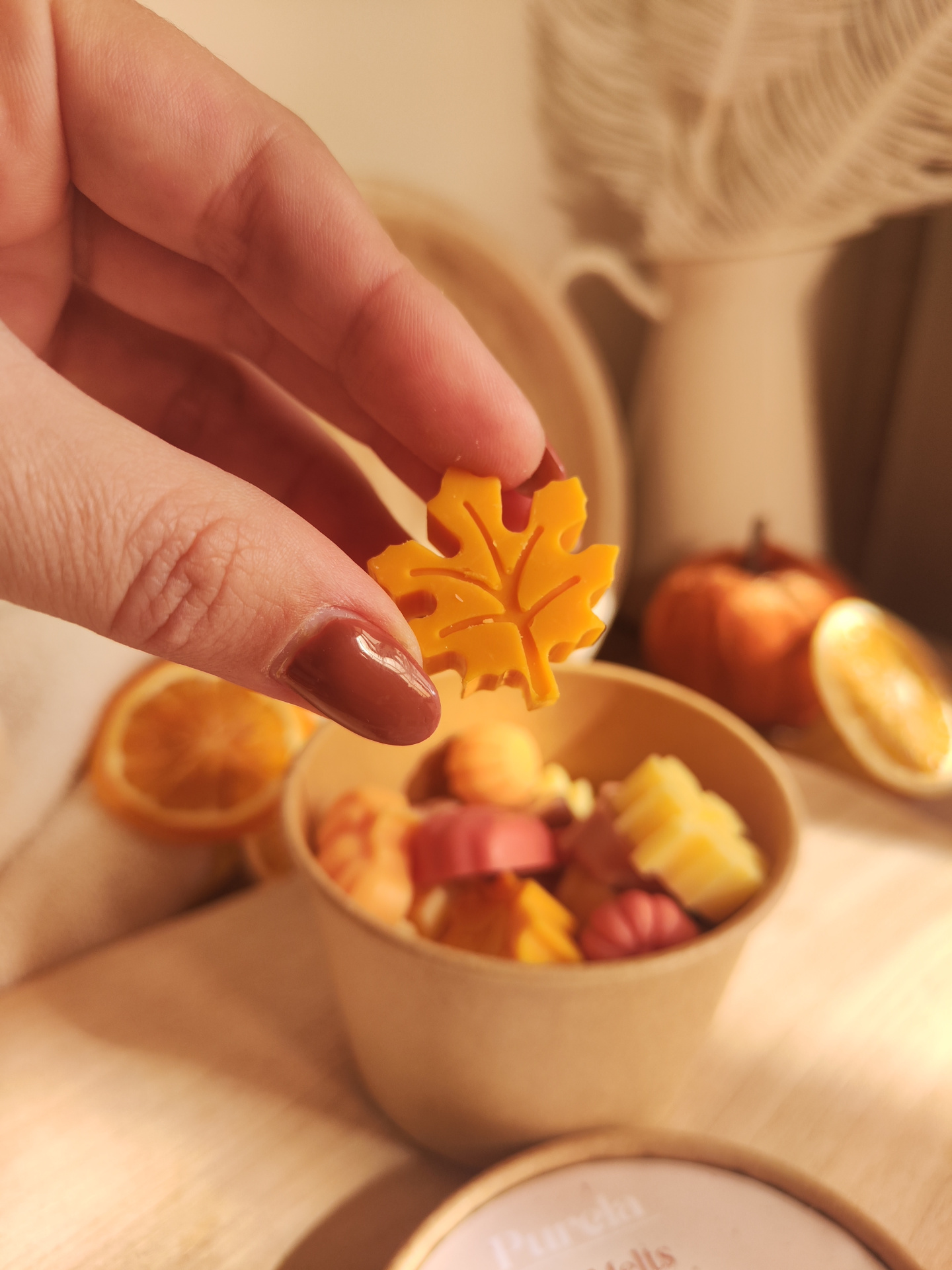 Wax Melt Laranja e Especiarias Mão segurando peça laranja em forma de folha com recipiente de papel cheio de decorações em forma de abóbora e folha no fundo