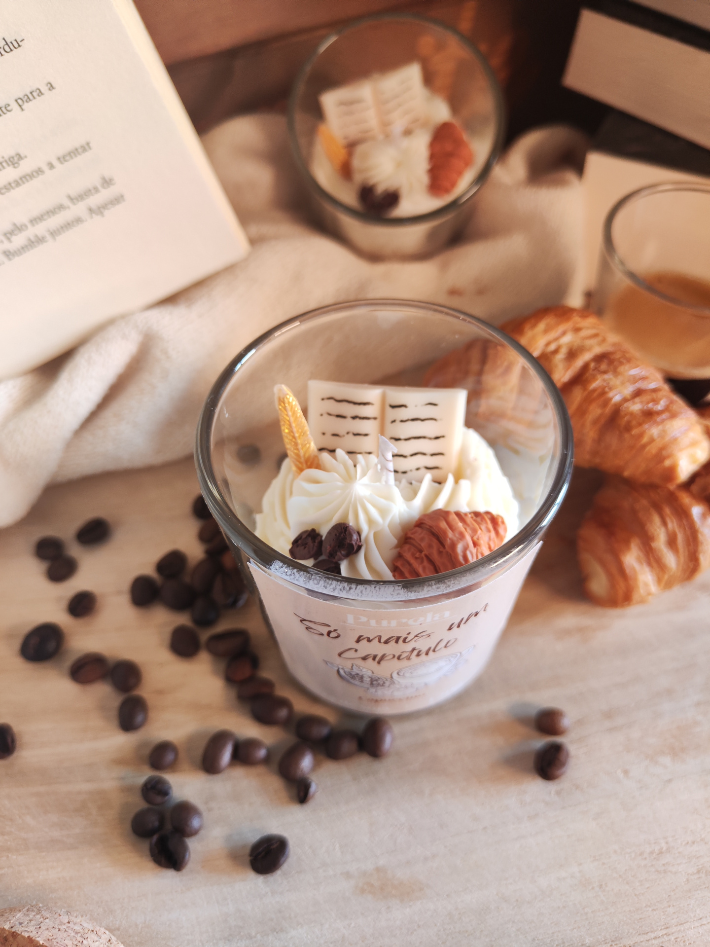 Vela Só mais um Capítulo Vela decorativa em copo de vidro com creme branco, decorada com pedaços e nozes, grãos de café ao redor, croissants e chávena de café ao fundo