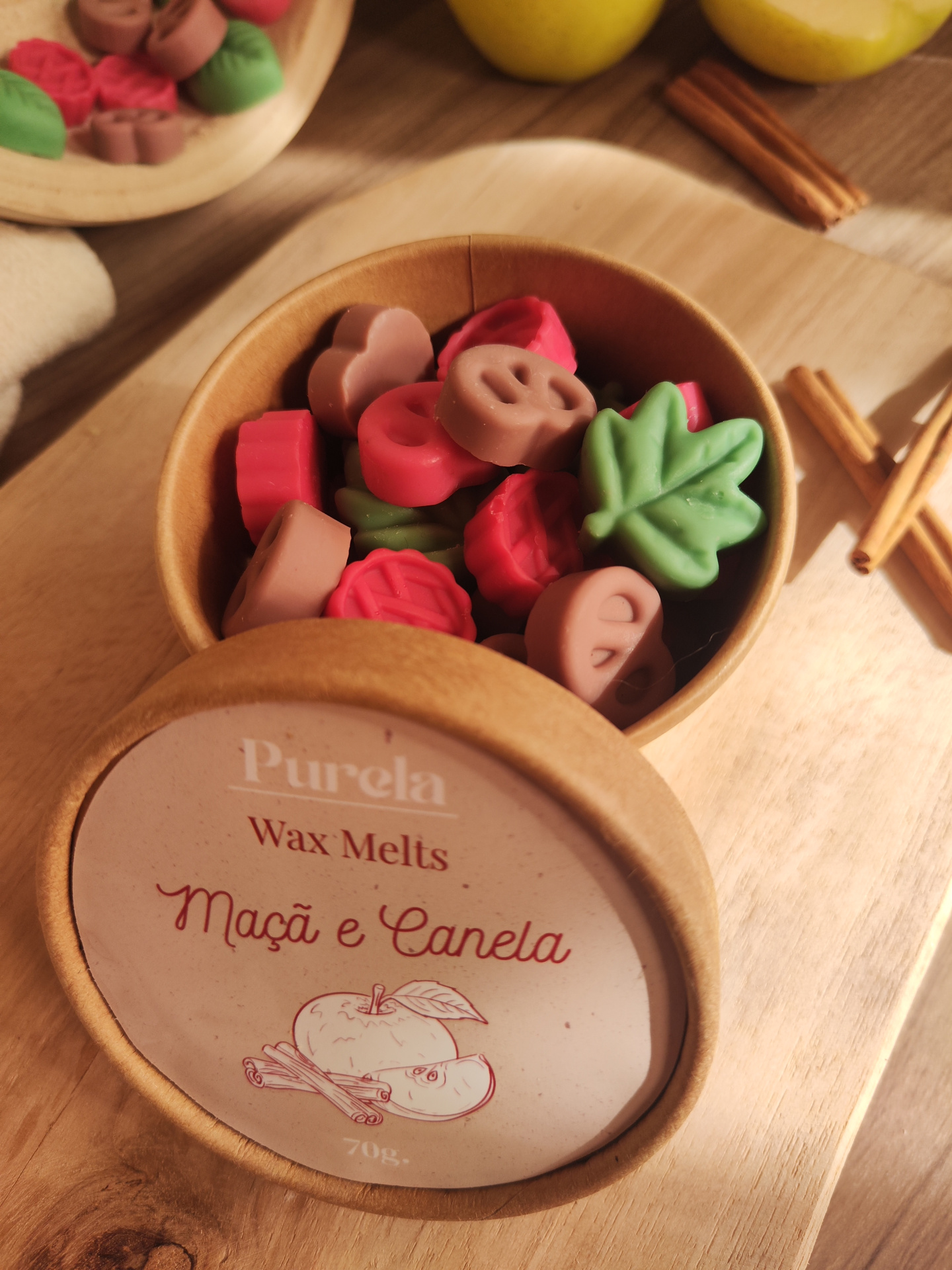 Wax Melt Maçã e Canela Recipiente com wax melts coloridos em forma de folhas e pretzels, etiqueta com texto Purela Wax Melts Maçã e Canela 70g sobre madeira