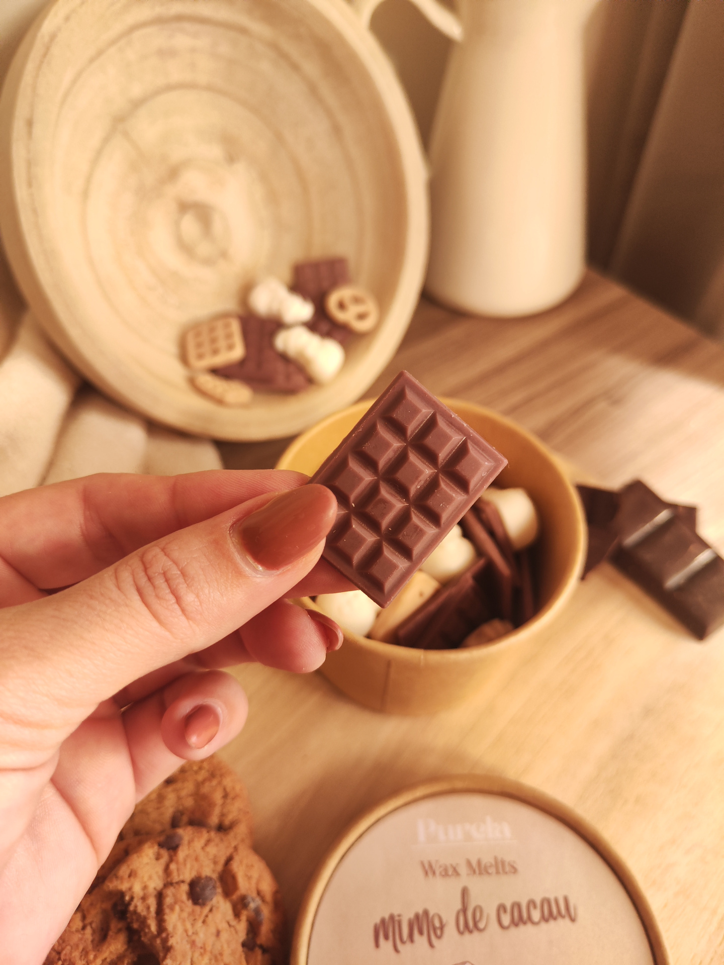 Wax Melt Mimo de Cacau Mão a segurar quadrado de cera marrom, cookies e caixa redonda com texto em cima de mesa de madeira