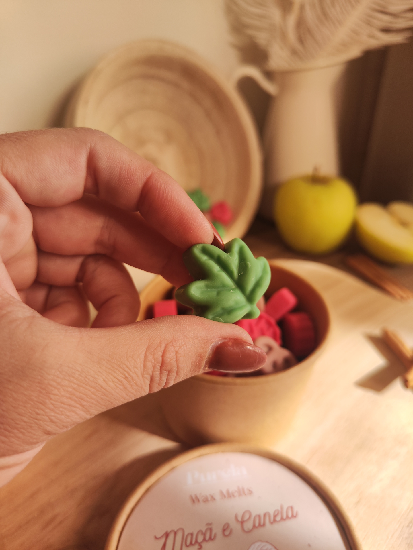 Wax Melt Maçã e Canela Mão segurando cera em forma de folha verde com recipiente de ceras coloridas e maçã amarela ao fundo