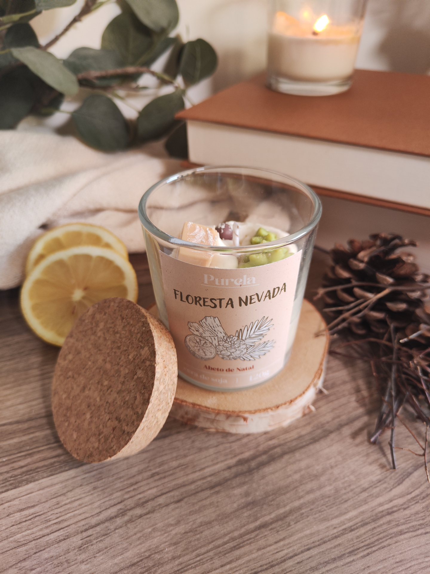 Vela Floresta Nevada Vela aromática em vidro com tampa de cortiça e etiqueta FLORESTA NEVADA, decorada com limão, folhas verdes, livro e vela ao fundo