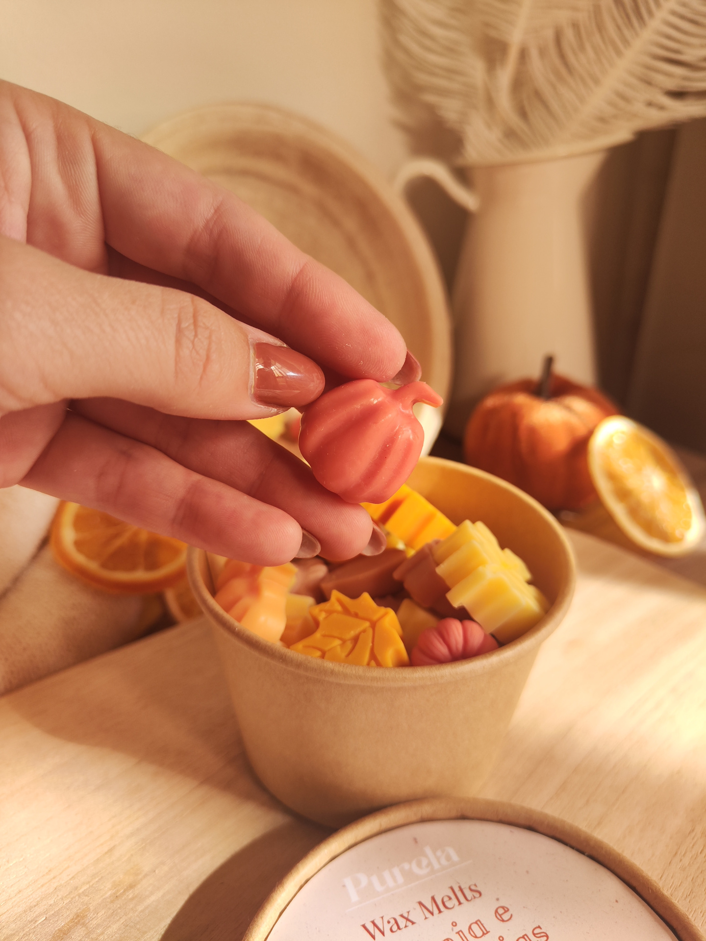 Wax Melt Laranja e Especiarias Mão segurando cera em forma de abóbora rosa com outras ceras coloridas em recipiente de papel castanho e fundo com decoração outonal.