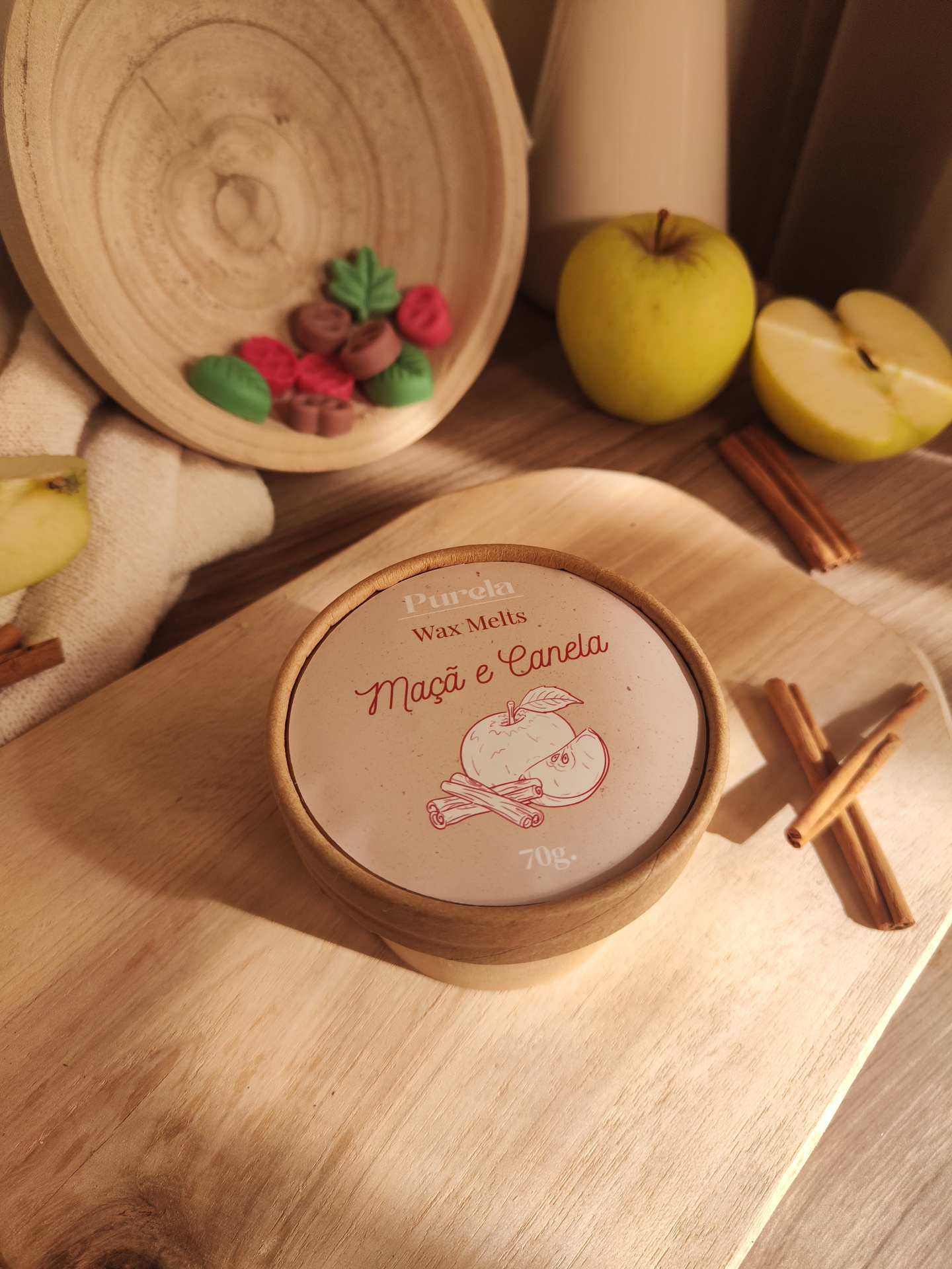 Wax Melt Maçã e Canela Caixa redonda de papel com label Purela Wax Melts Maçã e Canela 70g em mesa de madeira com paus de canela, maçãs e peça decorativa.
