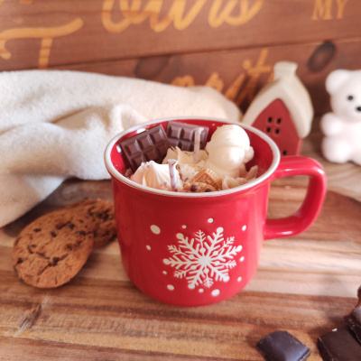 Caneca vermelha com floco de neve branco cheia de chantilly e chocolates, biscoitos e decoração natalícia ao fundo